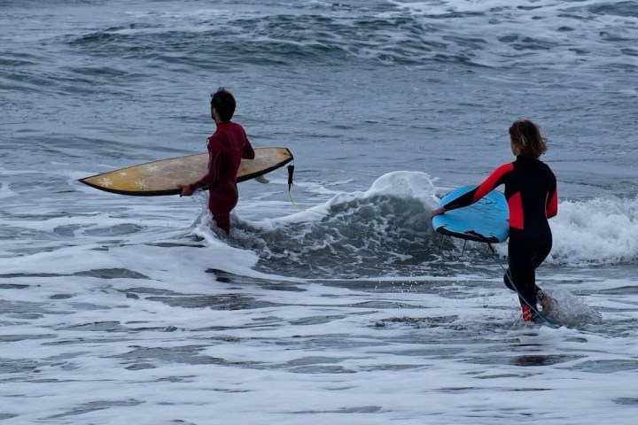 Two surfers in wetsuits enter the ocean with their boards on a cloudy day.