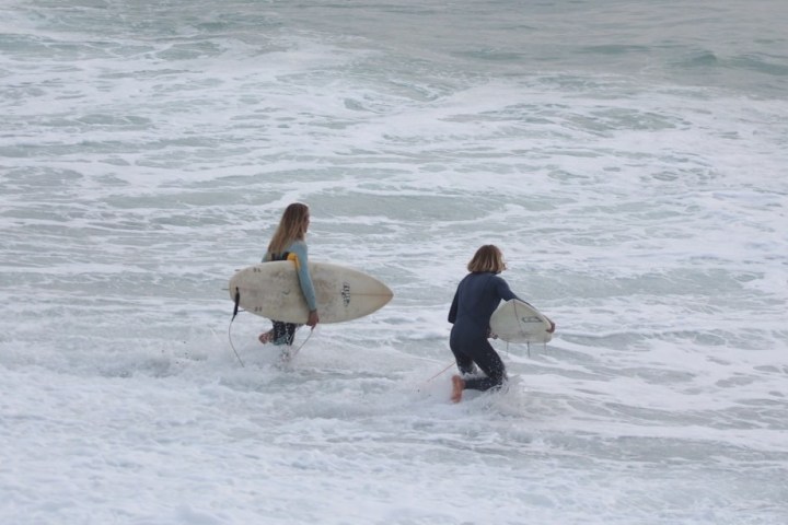 Two surfers in wetsuits walk into the ocean carrying surfboards amid white waves.