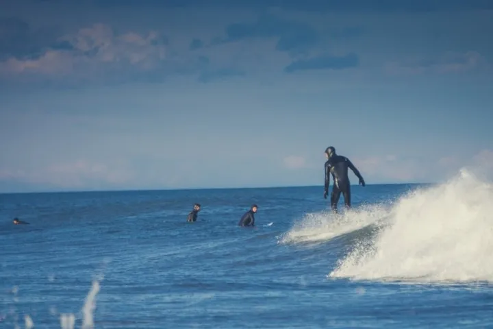 Surfer in a wetsuit rides a wave, with others watching in the ocean under a cloudy sky.