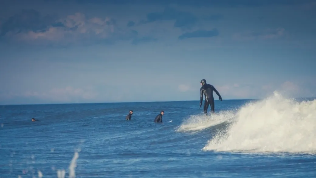 Surfer in a wetsuit rides a wave, with others watching in the ocean under a cloudy sky.