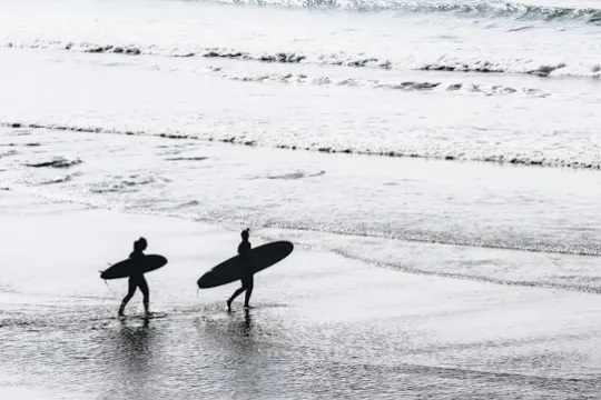 Two people walk on a beach carrying surfboards, ocean waves in the background.
