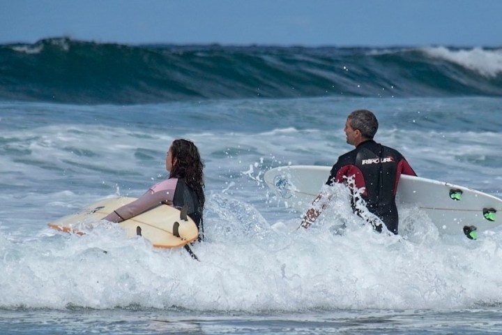 Two surfers with boards entering the ocean waves in wetsuits.