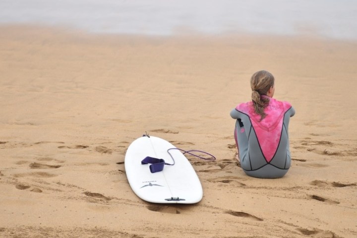 Person in wetsuit sitting on beach beside a surfboard, facing the sea.