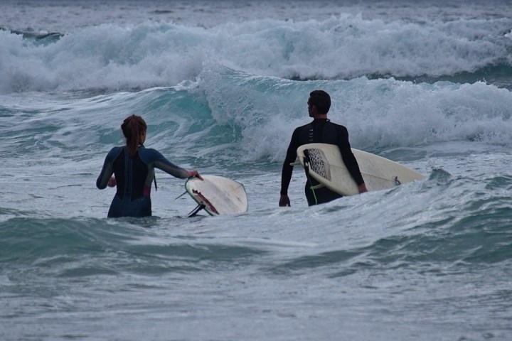 Two surfers in wetsuits carrying boards wade into ocean waves.