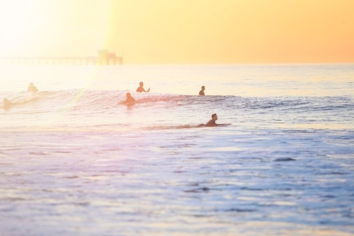 Surfers enjoy waves at sunset with a pier in the background and a bright, colorful sky.