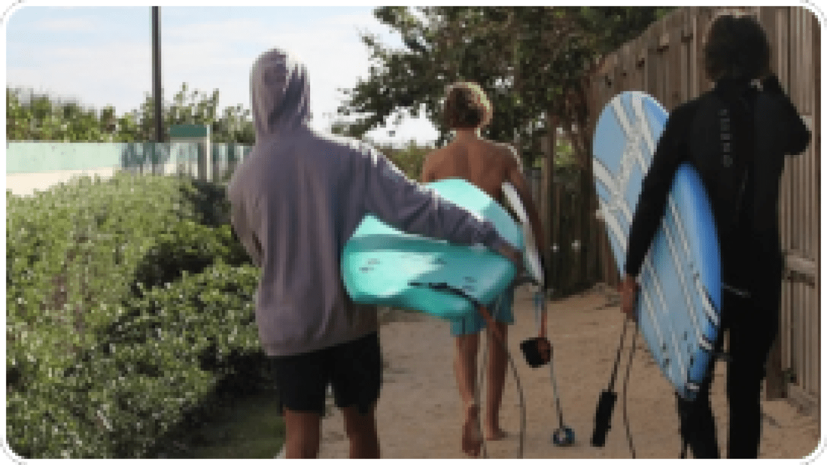 Three people walking with surfboards on a sandy path near bushes and a fence.