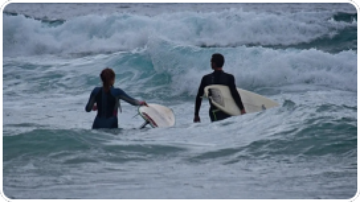 Two people in wetsuits carrying surfboards into the ocean waves.