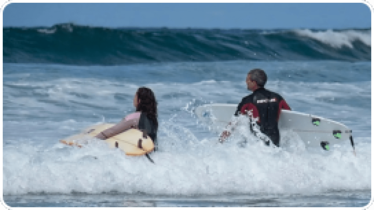 Two surfers in wetsuits carrying boards, wading through waves toward the ocean.
