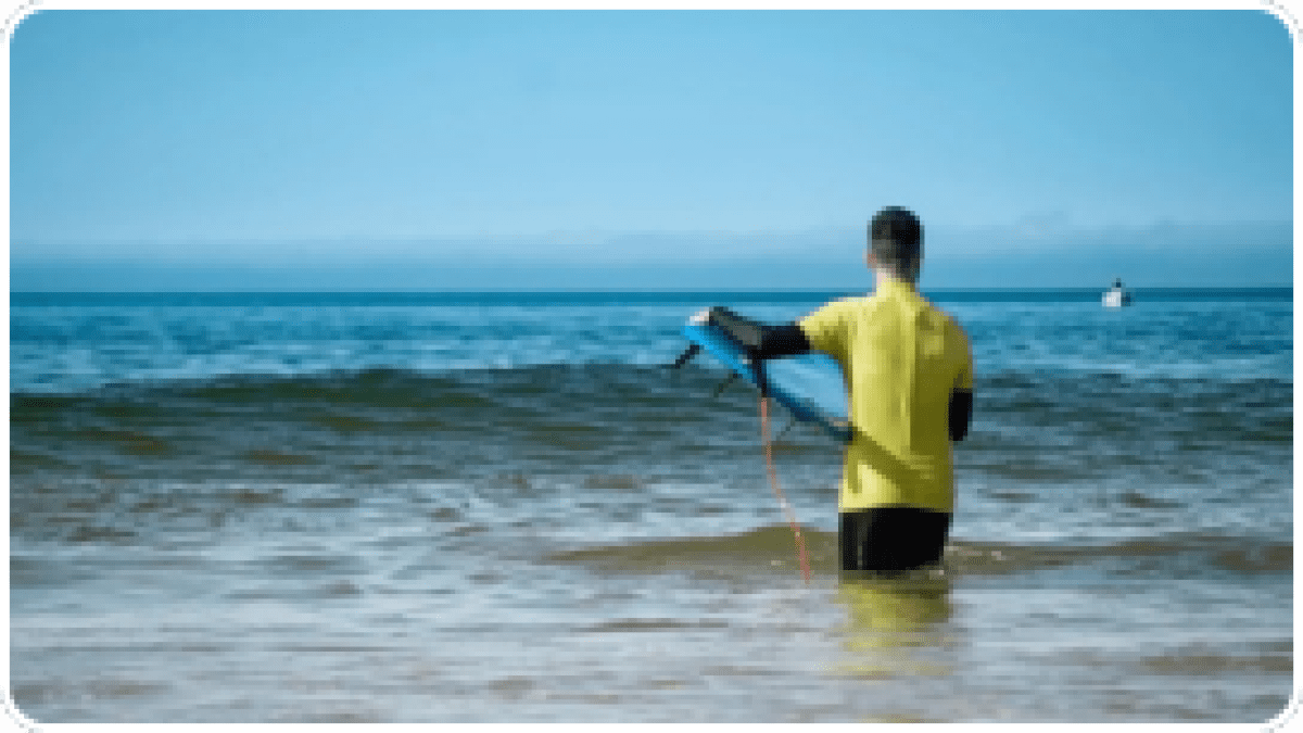 Person in yellow shirt holding a bodyboard, standing in shallow ocean water.