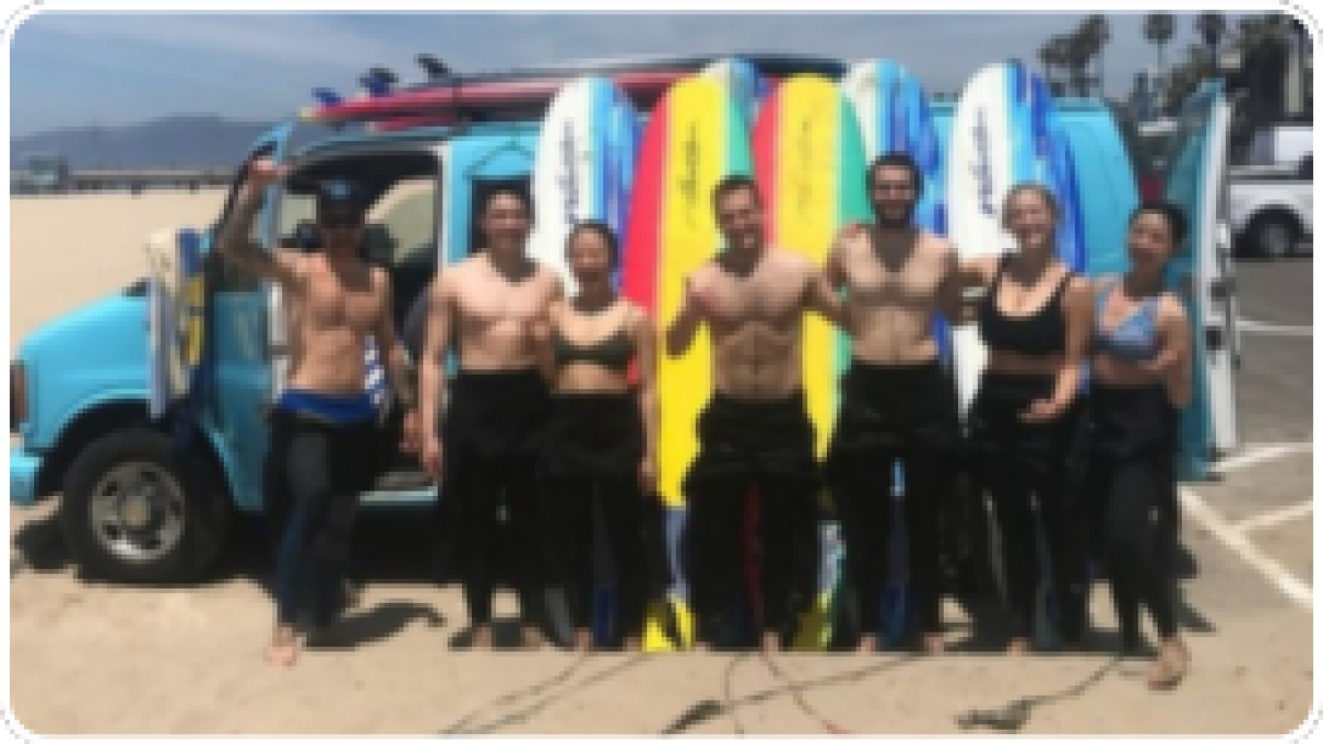Group of people in wetsuits holding surfboards by a blue van on a sunny beach.