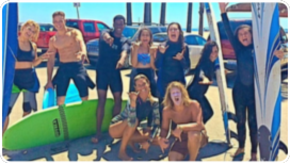 Group of smiling surfers with boards, posing happily near a beach with cars and palm trees in the background.