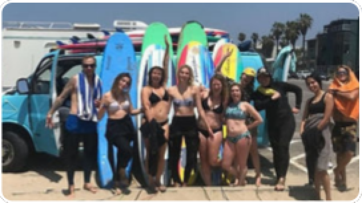 Group with surfboards poses by a blue van on a sunny beach day.
