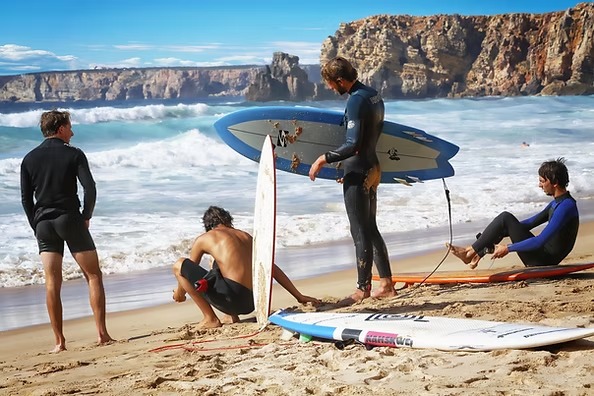 imgi_2_bbe0dc710b6b48b3baab18e69d564dd4 Surfers on a sandy beach with surfboards, rocky cliffs in the background.